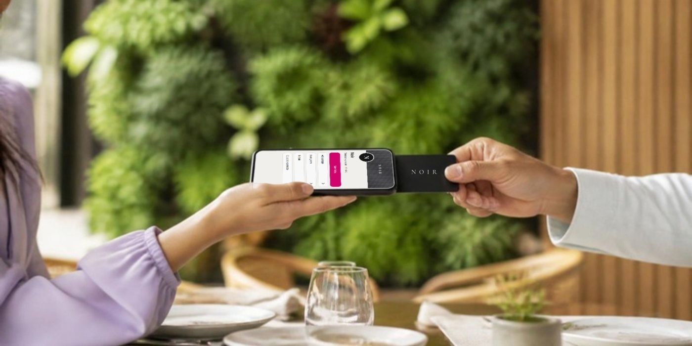 En la terraza de un restaurante, una persona acerca su celular a la NOIR card que le ofrece otra y se abre la página de Noir